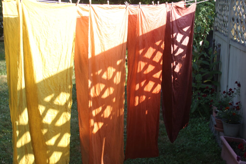 Yellow, orange, and red fabric glistens in the sun. The light on the yellow fabric in the foreground is glaring and makes a bright contrast with the shadows. Shadows from the fence make a pattern of diagonal lines. 