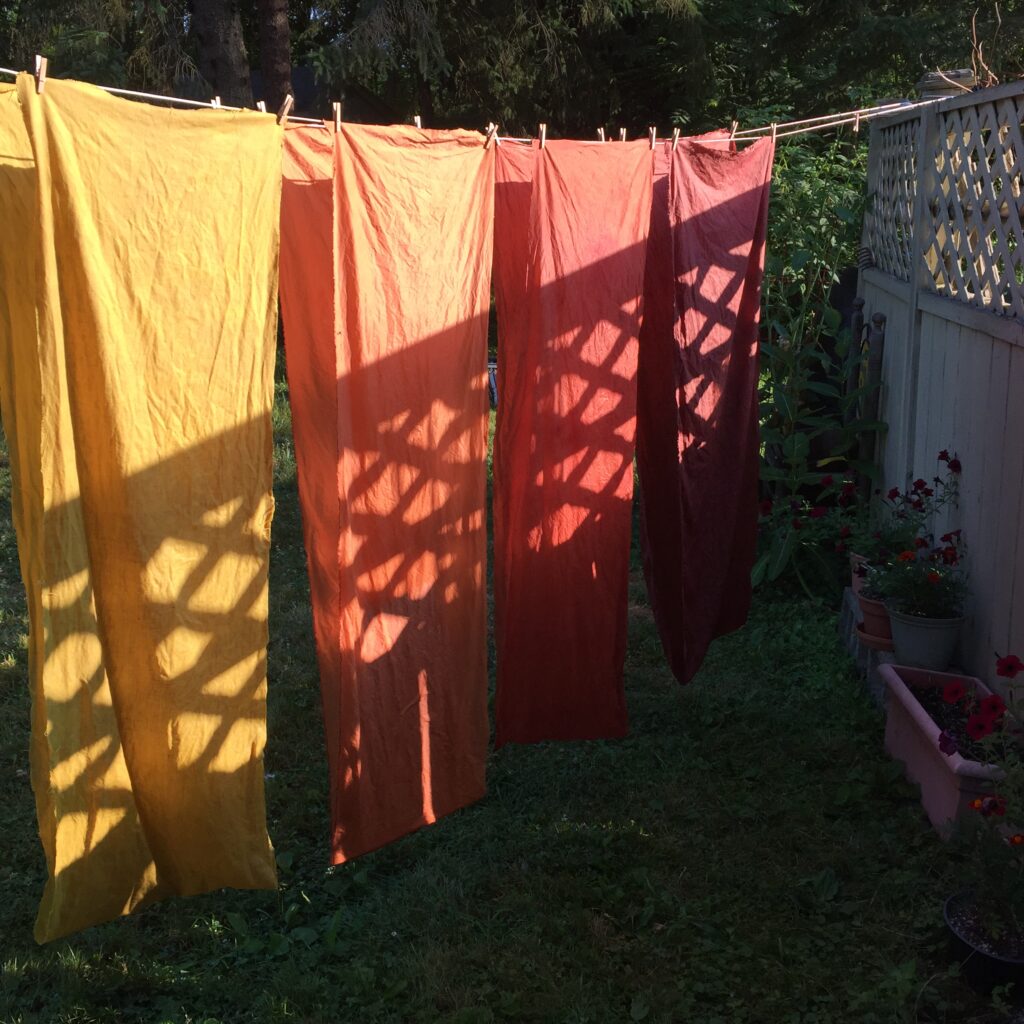Yellow, orange, and red fabric are drying in the early morning sun. Shadows from the fence make a pattern of diagonal lines.