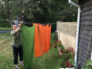 The author is smiling while she hangs green and orange cloth on a laundry line. Pots of red petunias are lined up at the base of a tan-colored fence.
