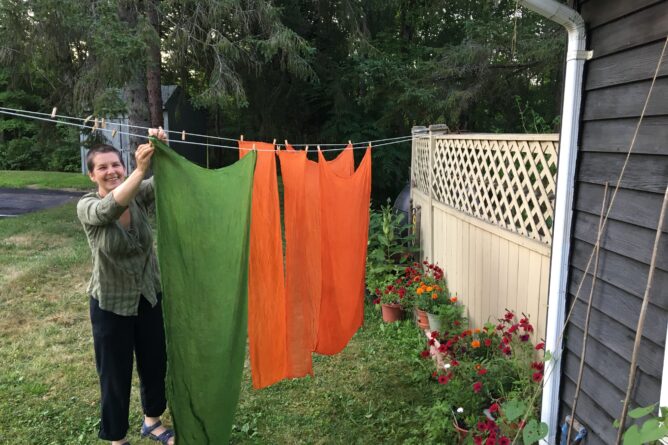 The author is smiling while she hangs green and orange cloth on a laundry line. Pots of red petunias are lined up at the base of a tan-colored fence.